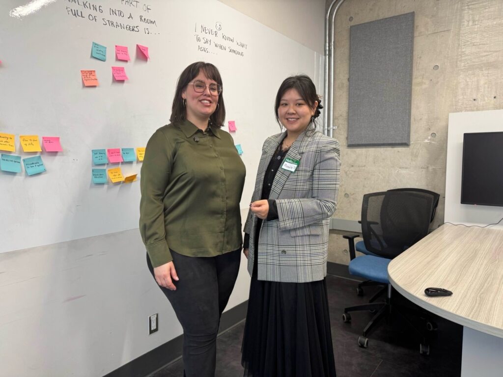 Boost Academy of Excellence CEO and another woman smile and stand together in front of a whiteboard covered in colorful sticky notes and handwritten prompts about networking. One wears a green shirt and glasses, the other a plaid blazer and name tag. The setting appears to be a workshop or event space.