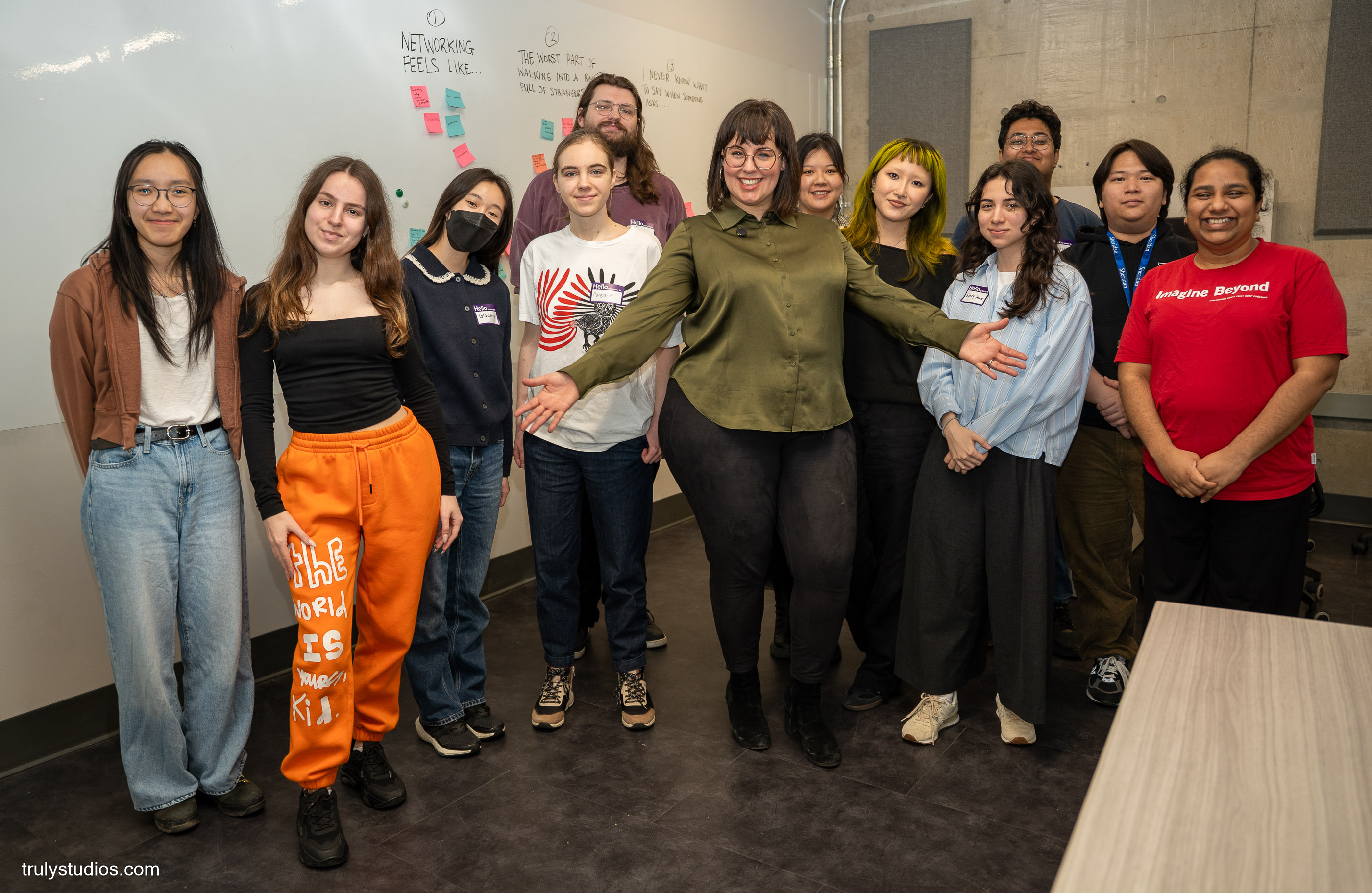 A group of approximately twelve young people posing together in front of a whiteboard covered in sticky notes and handwritten prompts about networking. Several participants wear name tags. One person in the center, wearing a green shirt and glasses, spreads her arms wide to embrace the group. The setting appears to be a workshop or classroom event.