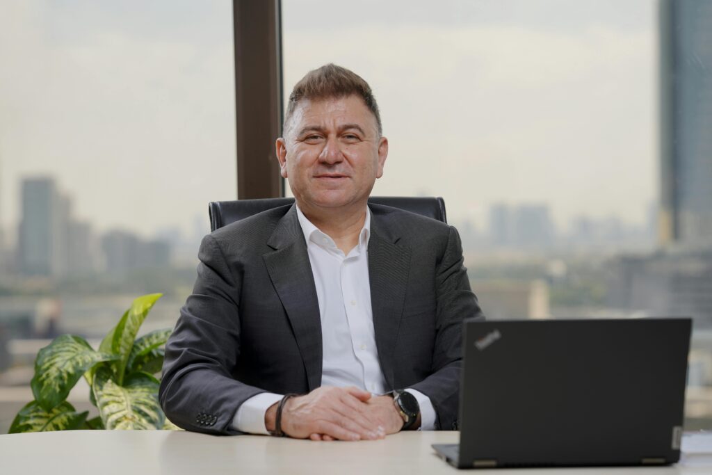 A man in a dark blazer and white shirt sits at a desk with hands folded, smiling confidently. A laptop and small plant are on the desk beside him. A large window behind him reveals a blurred city skyline.
