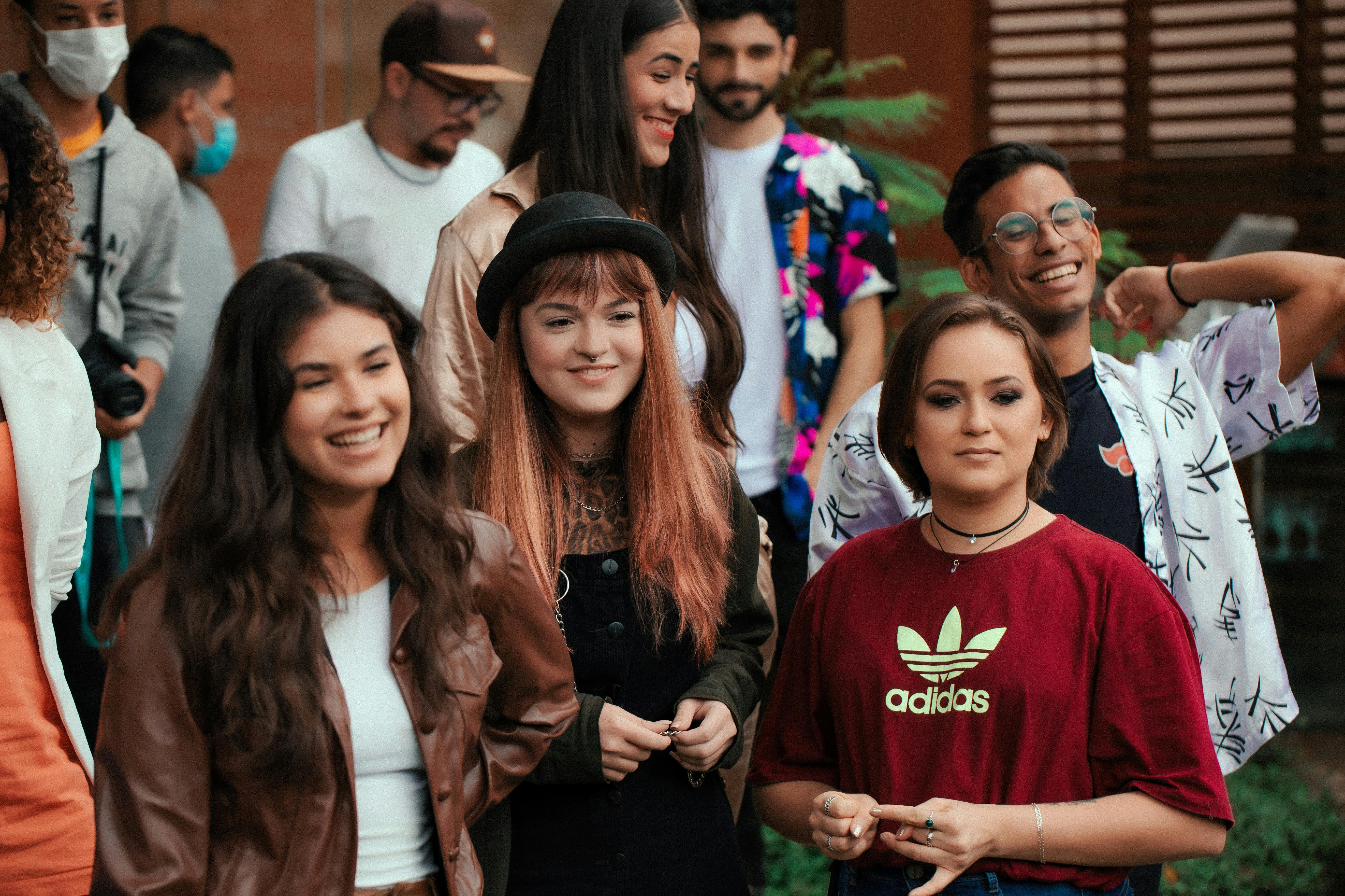 A group of young adults gathered outdoors, smiling and socializing. Three women stand in the foreground — one wearing a brown jacket, one in a black outfit with a hat and red hair, and one in a red Adidas t-shirt. Several people are visible in the background, one wearing a face mask.